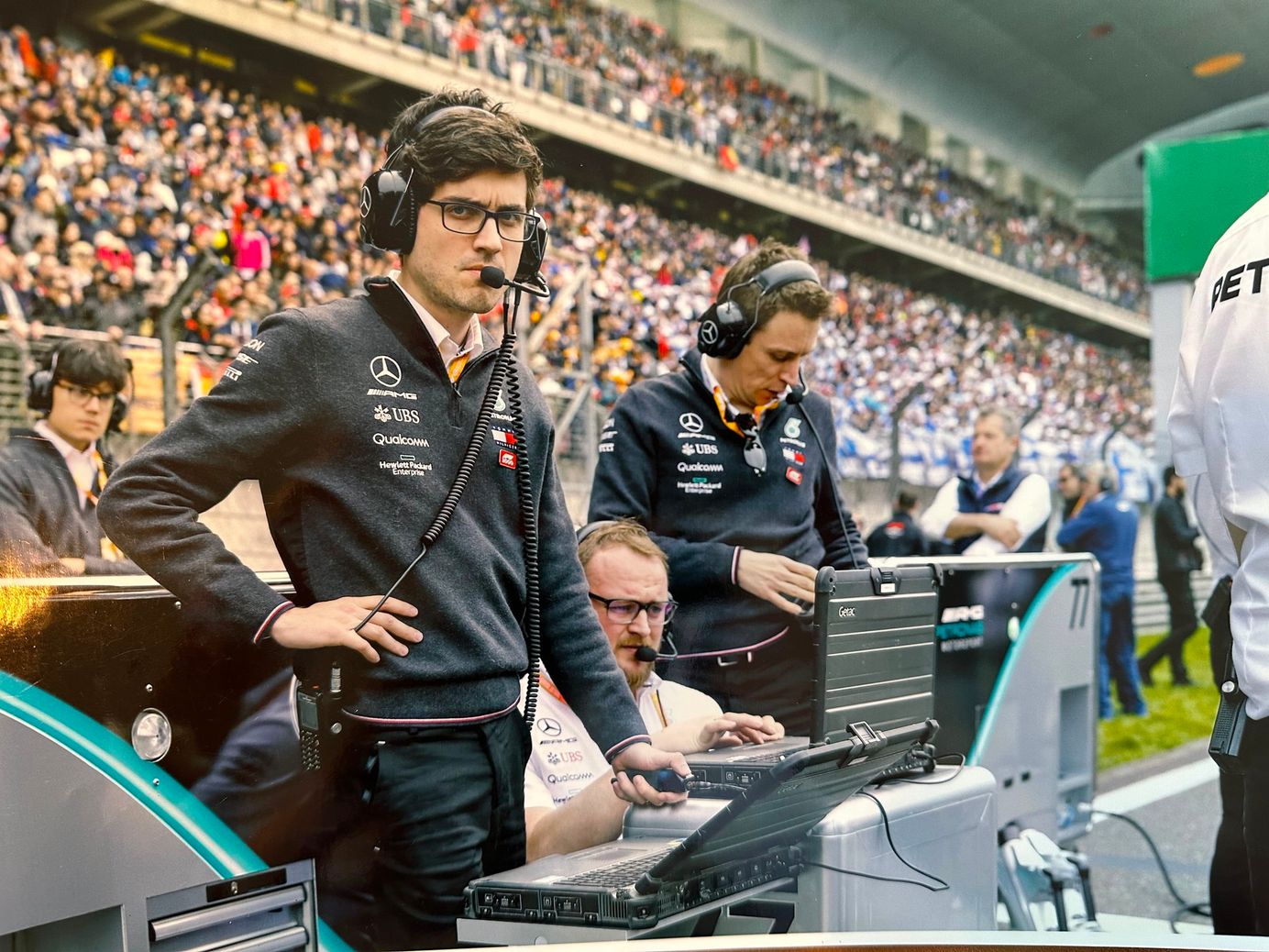 Luiz Oliveira at the Mercedes pit wall in team kit and headset, a grandstand behind