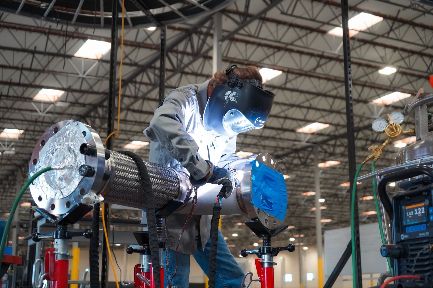 A technician in a welding mask welds a flanged pipe assembly on the shop floor