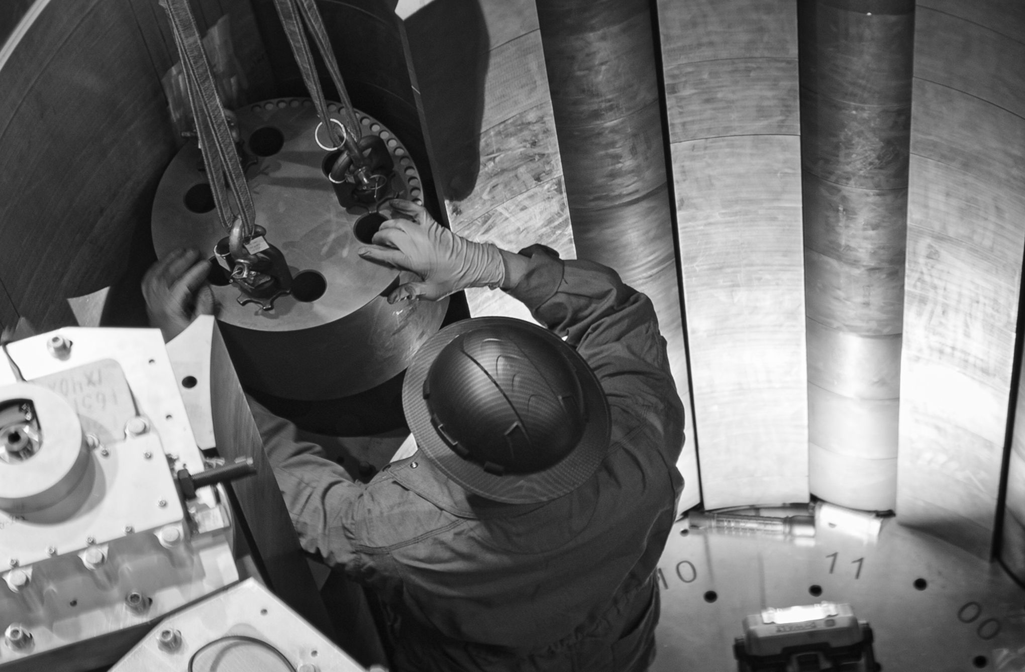 Black-and-white photo from above of a technician in a hard hat fitting lifting slings to a reactor component