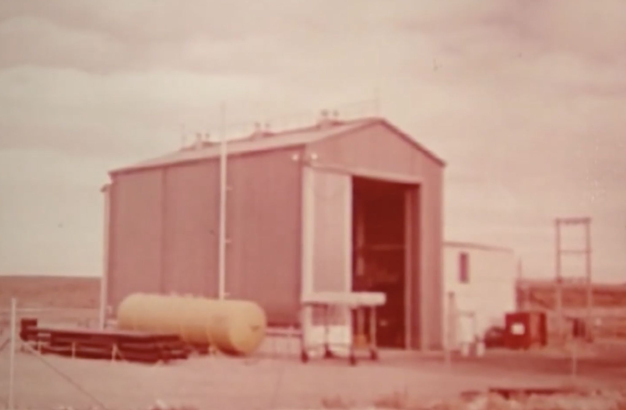 Archival photo of a small metal test building and fuel tank on open, flat ground